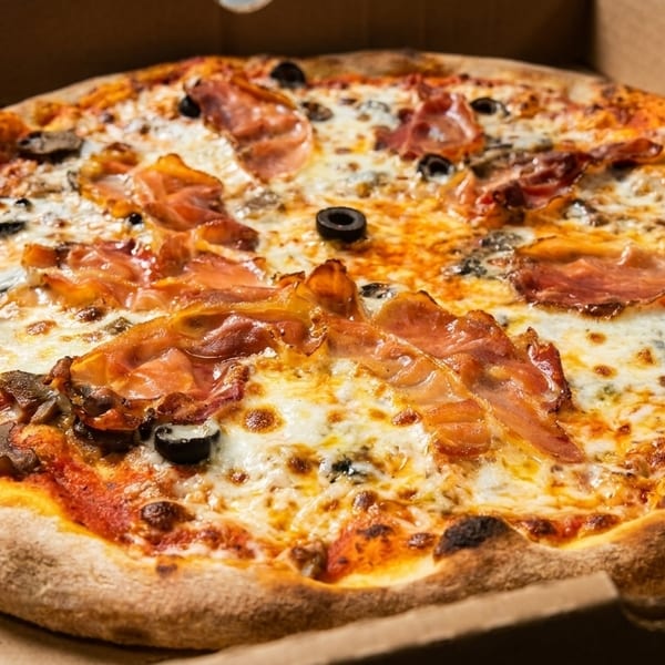 A freshly baked pizza with prosciutto, mushrooms, and olives in a cardboard delivery box.